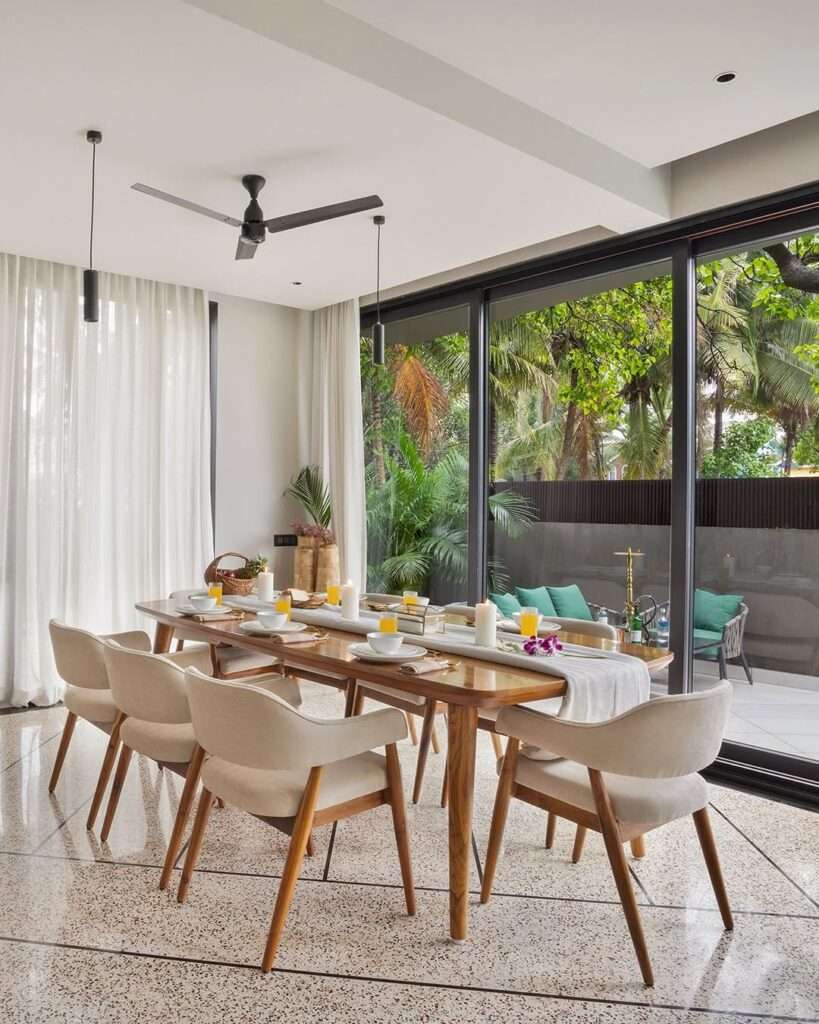 Modern dining space with wooden table, upholstered chairs, large glass windows, and indoor-outdoor connection designed by Amartya Architects for elegant residential interiors.